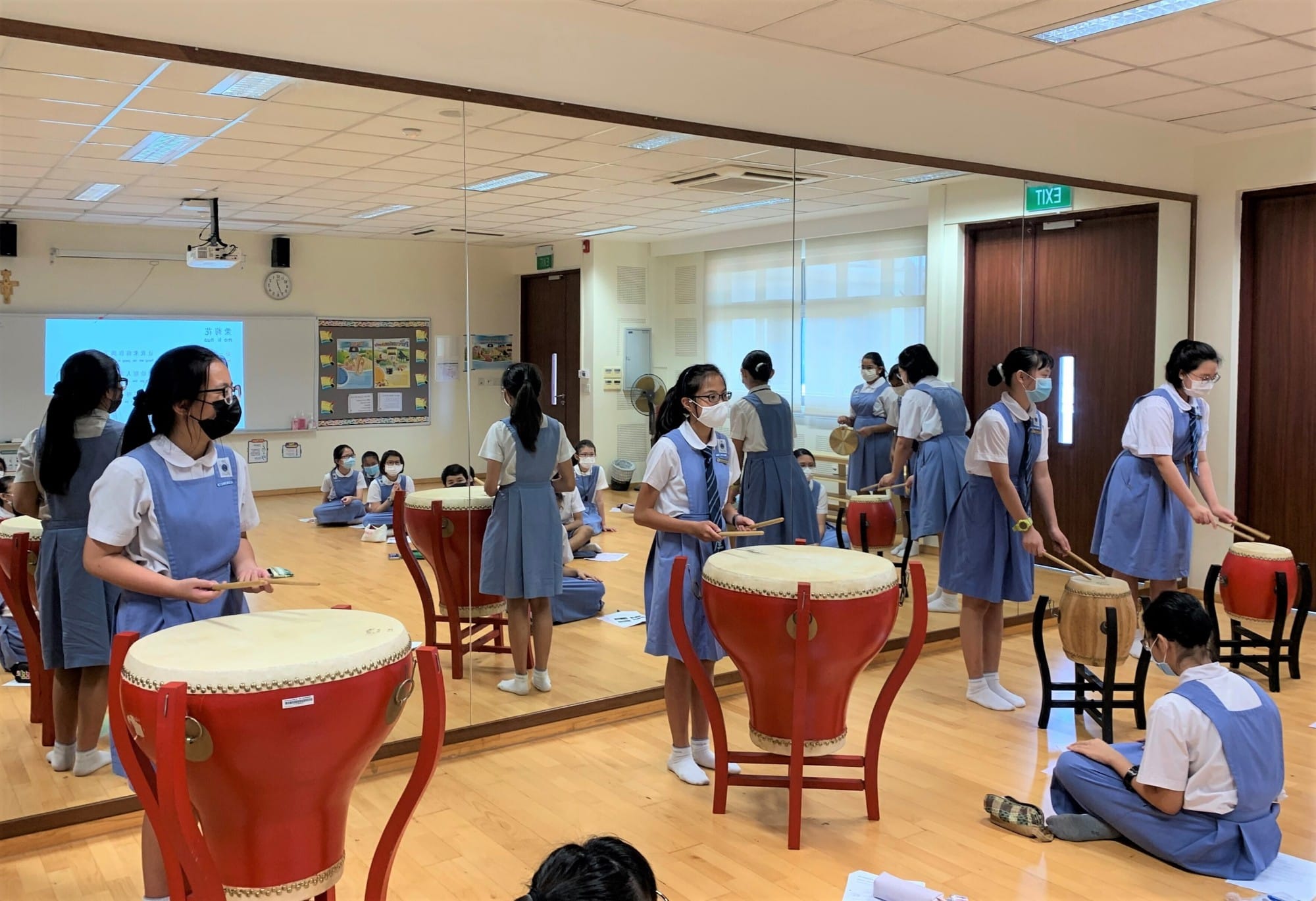 Masked schoolgirls in blue uniforms play large red Chinese drums with drumsticks in a mirrored classroom.
